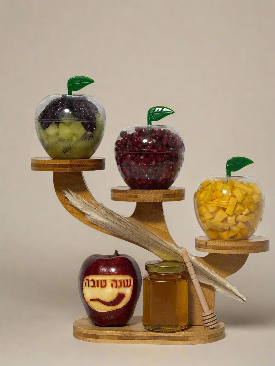 Three glass apples with fruits, a honey jar, and a lulav on a wooden stand against a white background.